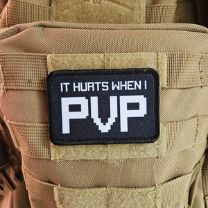 May include: A tan tactical backpack with a black and white patch. The patch says "IT HURTS WHEN I PVP". The rectangular patch is attached to the front of the backpack. The backpack has multiple straps and a velcro patch.