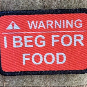 Può includere: Toppa di avvertimento rossa con bordo nero e testo bianco che recita "WARNING I BEG FOR FOOD" con un triangolo bianco con un punto esclamativo all'interno.