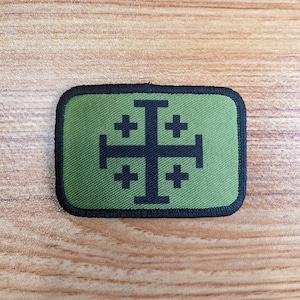 Può includere: Una toppa ricamata verde con una croce nera e quattro croci più piccole intorno. La toppa ha un bordo nero.