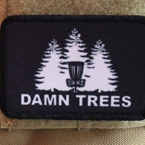 May include: Black rectangular patch featuring a white graphic of three pine trees and a disc golf basket. The text "DAMN TREES" is printed in white below the graphic. The patch is attached to a tan fabric.