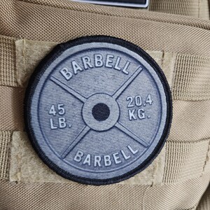 May include: A round, gray patch with the word "BARBELL" arched at the top and bottom. The center features a weight plate design with "45 LB." and "20.4 KG." printed on it. The patch is attached to a tan fabric.