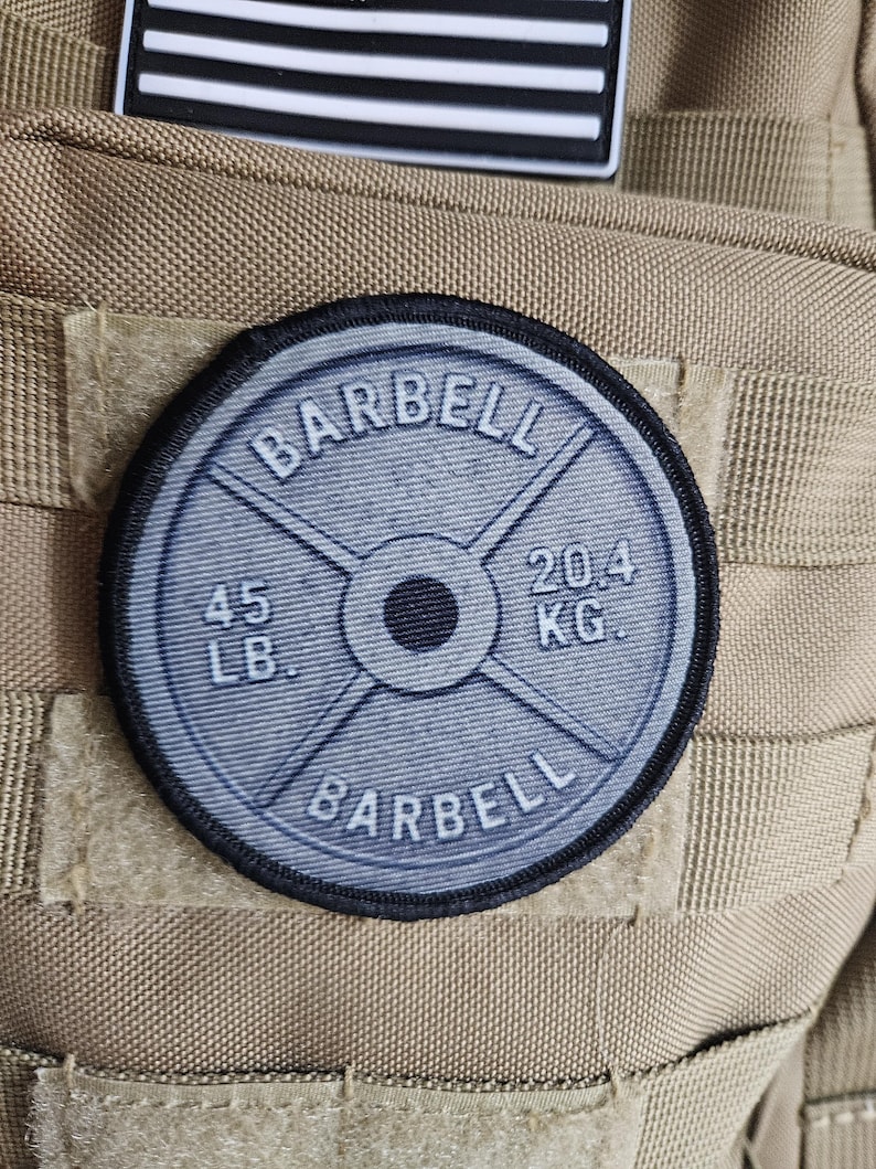 May include: A round, gray patch with the word "BARBELL" arched at the top and bottom. The center features a weight plate design with "45 LB." and "20.4 KG." printed on it. The patch is attached to a tan fabric.