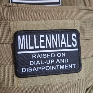 Könnte beinhalten: Ein schwarzer rechteckiger Aufnäher mit dem Wort "MILLENNIALS" in weißen Blockbuchstaben über dem Text "RAISED ON DIAL-UP AND DISAPPOINTMENT". Ein amerikanischer Flaggenaufnäher befindet sich über dem Hauptaufnäher. Die Aufnäher befinden sich auf einem beigefarbenen Stoff.