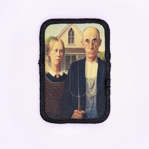 May include: A black and white embroidered patch depicting a painting of a man and woman standing in front of a house. The man is wearing a black suit and holding a pitchfork. The woman is wearing a black dress. The house is white with a black roof.