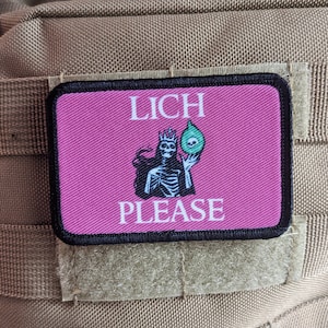 Może przedstawiać: Różowa łatka z czarnym obszyciem i napisem "LICH PLEASE" w kolorze białym. W środku przedstawiono szkielet w koronie trzymający zieloną kulę.