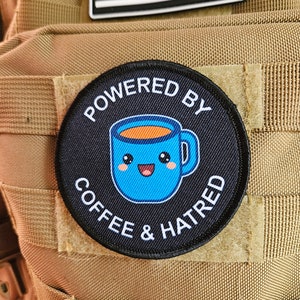 Puede incluir: Un parche blanco y negro con el texto "Powered by Coffee & Hatred" y una taza de café azul de dibujos animados con una cara sonriente.
