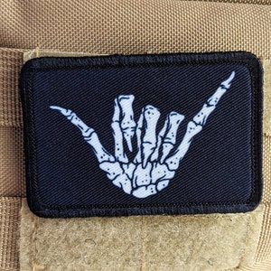 May include: A black patch with a white skeleton hand making the shaka sign. The patch is sewn onto a brown fabric background.