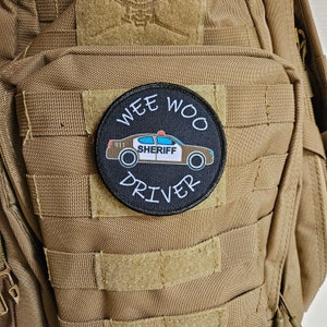 Puede incluir: Un parche blanco y negro con las palabras "Wee Woo Driver" y un coche de policía de dibujos animados con la palabra "Sheriff" en el lateral. El parche está sujeto a una mochila táctica marrón.