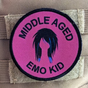 May include: A pink and black round patch with the text "Middle Aged Emo Kid" and a black silhouette of a person with blue hair.