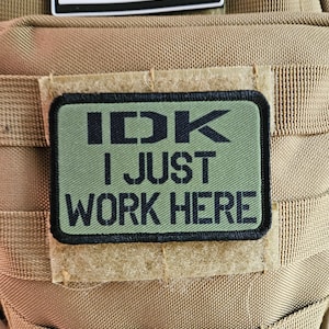 May include: A green patch with black text that reads "IDK I JUST WORK HERE" attached to a brown tactical vest.