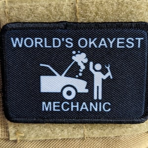May include: A black patch with white text that reads "WORLD'S OKAYEST MECHANIC". A white cartoon of a car with its hood open and smoke coming out of it is next to a white cartoon of a person holding a wrench.