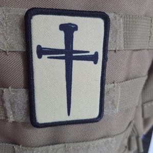 May include: A rectangular patch with a cream-colored background and a dark navy blue border. The center features a graphic design of two crossed iron nails forming a cross shape, rendered in the same dark navy blue color as the border.