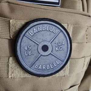 May include: A round patch featuring a barbell weight plate design. The patch is gray with black trim and displays the word "BARBELL" and weight measurements of "45 LB." and "20.4 KG.". The patch is attached to a tan tactical vest.