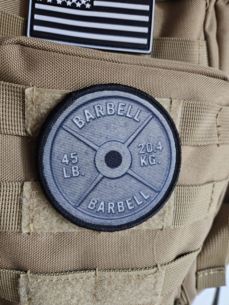 May include: A round patch featuring a barbell weight plate design. The patch is gray with black trim and displays the word "BARBELL" and weight measurements of "45 LB." and "20.4 KG.". The patch is attached to a tan tactical vest.
