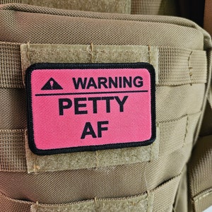 May include: A pink patch with black trim and text that reads "WARNING PETTY AF" attached to a brown tactical backpack.