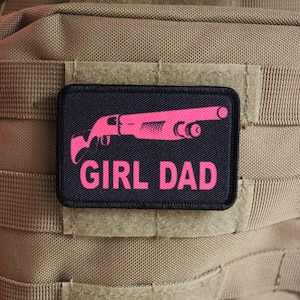 以下が含まれることがあります： 「GIRL DAD」というピンクの文字と、ピンクのショットガンのシルエットが描かれた黒いパッチ