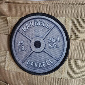 May include: A circular patch featuring a barbell weight plate design. The patch is gray with black text that reads "BARBELL", "45 LB", and "20.4 KG". The patch is attached to a tan fabric.