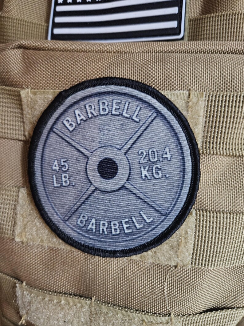 May include: A circular patch featuring a barbell weight plate design. The patch is gray with black text that reads "BARBELL", "45 LB", and "20.4 KG". The patch is attached to a tan fabric.