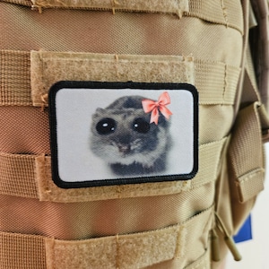 May include: A brown tactical backpack with a black patch featuring a cartoon animal with a pink bow. The patch is attached to the front of the backpack with velcro.