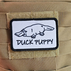 Puede incluir: Un parche blanco con ribete negro presenta un dibujo en blanco y negro de un ornitorrinco con el texto "Duck Puppy" debajo.