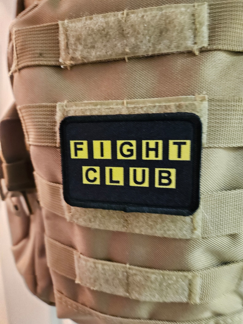 Peut inclure: Un gilet tactique beige avec un patch noir qui dit "FIGHT CLUB" en lettres jaunes.