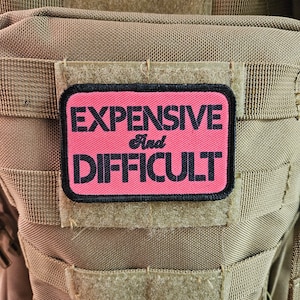 Puede incluir: Un parche rosa con texto negro que dice "EXPENSIVE and DIFFICULT" está unido a un chaleco táctico marrón.