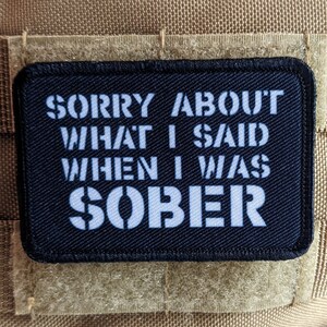 以下が含まれることがあります： 「SORRY ABOUT WHAT I SAID WHEN I WAS SOBER」と書かれた白いステンシル文字が特徴の、長方形の黒いパッチ。 パッチは、ベージュ色の布地の背景に配置されています。