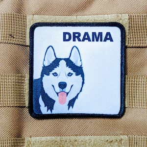 May include: A white patch with black trim features a cartoon husky dog with blue eyes and a pink tongue. The text "DRAMA" is printed in black at the top of the patch.