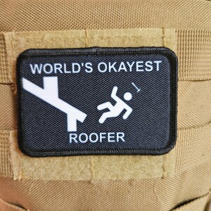May include: Black patch with white text and graphic. The text reads "World's Okayest  Roofer". The graphic is a stick figure falling off a roof.