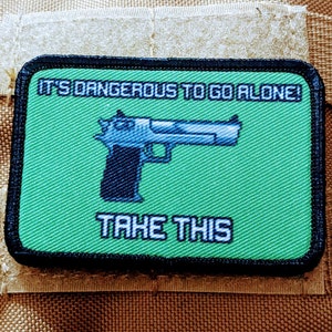May include: A rectangular patch with a green background and black border. The patch features a pixelated image of a handgun and the text "IT'S DANGEROUS TO GO ALONE! TAKE THIS" in white.