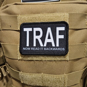 Puede incluir: Un parche negro con texto blanco que dice "TRAF" y "NOW READ IT BACKWARDS" sobre un fondo marrón.