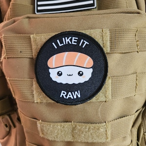 Puede incluir: Un parche blanco y negro con un rollo de sushi de dibujos animados y el texto "I LIKE IT RAW" en una mochila táctica de color marrón.