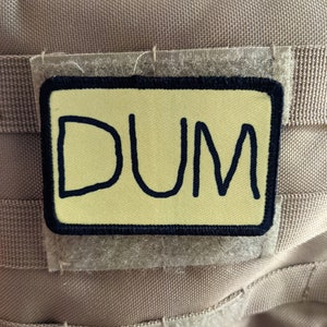 Può includere: Una toppa rettangolare con la scritta "DUM" in lettere blu scuro, in stile manoscritto, su sfondo giallo chiaro. La toppa ha un bordo nero ed è attaccata a un tessuto beige con chiusure a strappo.