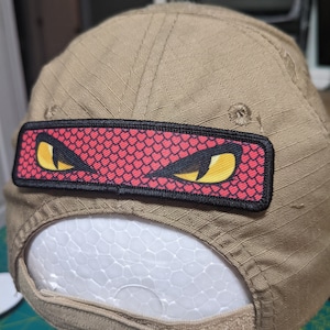 Pode incluir: Um boné de beisebol bege com um remendo preto e vermelho com um par de olhos amarelos. O remendo tem um fundo vermelho com contorno preto e um padrão de pequenos corações vermelhos.