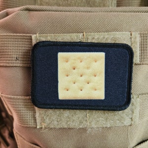 May include: A black patch with a white square-shaped cracker in the center. The cracker has a textured surface and is surrounded by a black border.