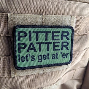 May include: Green and black patch with the text "Pitter Patter let's get at 'er" on a tan fabric background.