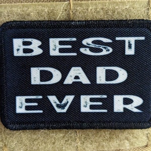 Può includere: Toppa rettangolare nera con le parole "BEST DAD EVER" in lettere bianche invecchiate. La toppa è bordata con cuciture nere ed è posta su uno sfondo di tessuto color cachi.
