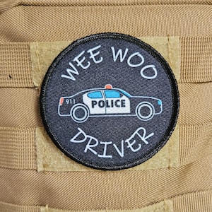 Może przedstawiać: Okrągła łatka w kolorze czarnym i białym z napisem "WEE WOO DRIVER" wokół krawędzi. W środku łatki znajduje się kreskówkowy radiowóz z napisem "POLICE" i "911".
