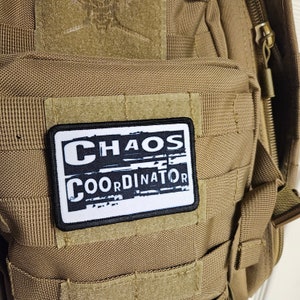 Peut inclure: Un sac &agrave; dos tactique beige avec un patch noir et blanc qui indique "CHAOS COORDINATOR".