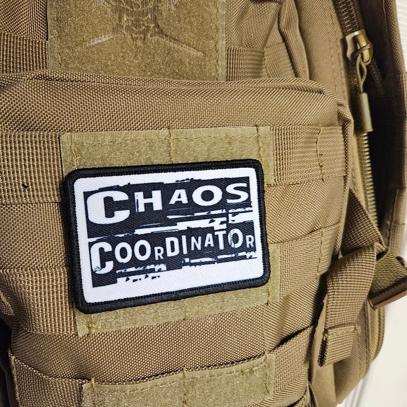 Peut inclure: Un sac &agrave; dos tactique beige avec un patch noir et blanc qui indique "CHAOS COORDINATOR".