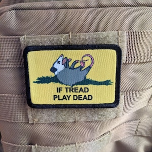 Pode incluir: Um remendo amarelo e preto com um opossum de desenho animado nele. O opossum está deitado de costas com as pernas no ar. O texto no remendo diz "If Tread Play Dead".