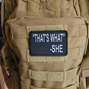 Pode incluir: Um remendo preto com texto branco que diz "THAT'S WHAT" -SHE. O remendo está preso a um colete tático bege.