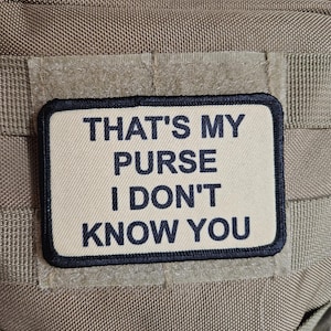 Pode incluir: Patch retangular bege com borda preta e o texto preto: "THAT'S MY PURSE I DON'T KNOW YOU". O patch está preso a um tecido bege com fecho de velcro.
