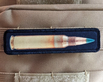 May include: A black patch with a white and gold bullet with a blue tip. The patch is sewn onto a brown fabric background.