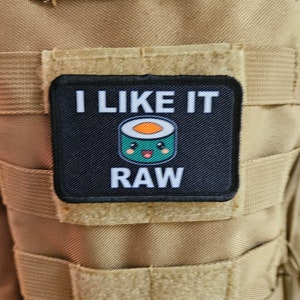 Puede incluir: Un parche blanco y negro con el texto "I LIKE IT RAW" y un rollo de sushi de dibujos animados con una cara sonriente.