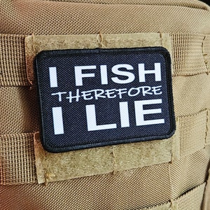 Puede incluir: Un parche negro con texto blanco que dice "I FISH THEREFORE I LIE" está unido a un chaleco táctico marrón.