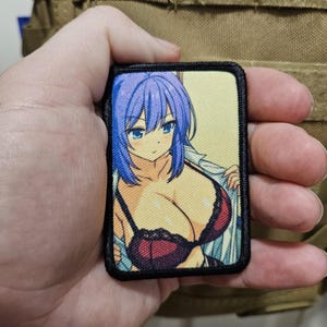 Peut inclure: Un patch rectangulaire avec une illustration de style anime d'une femme aux cheveux bleus et un soutien-gorge rouge. Le patch a une bordure noire et est tenu sur un fond beige.
