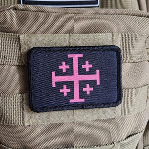 May include: A black rectangular patch featuring a pink Jerusalem cross design. The patch is affixed to a tan tactical backpack. Above the patch is a black and white American flag patch.