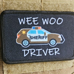 May include: Black patch with white text that reads "WEE WOO DRIVER". A cartoon image of a brown sheriff's car with a blue light on top is in the center of the patch.