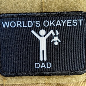 以下が含まれることがあります： 白文字で「WORLD'S OKAYEST DAD」と書かれた黒いパッチ。白い棒人間が、もう1人の小さな棒人間を逆さまに抱えている。